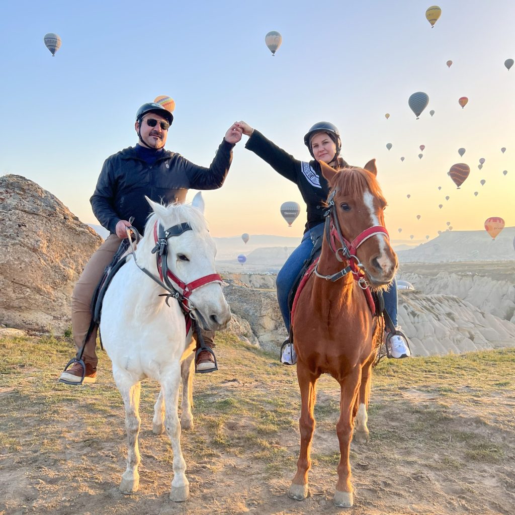 Riders on horseback during sunrise in Cappadocia with dozens of hot air balloons flying in the sky.